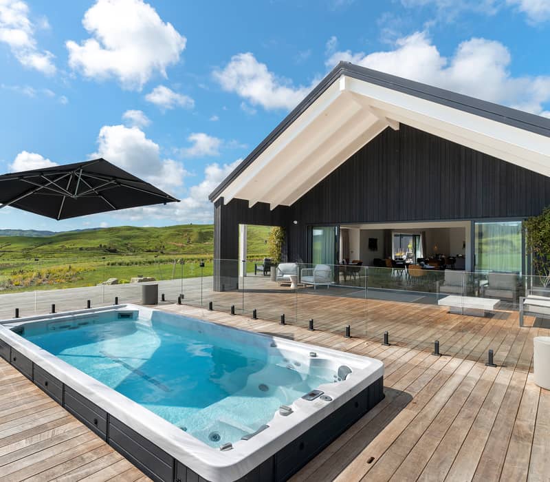 A luxury swim spa on a wooden deck outside a modern black barn-style lodge with green hills in the background Credit: Capstone Hotels