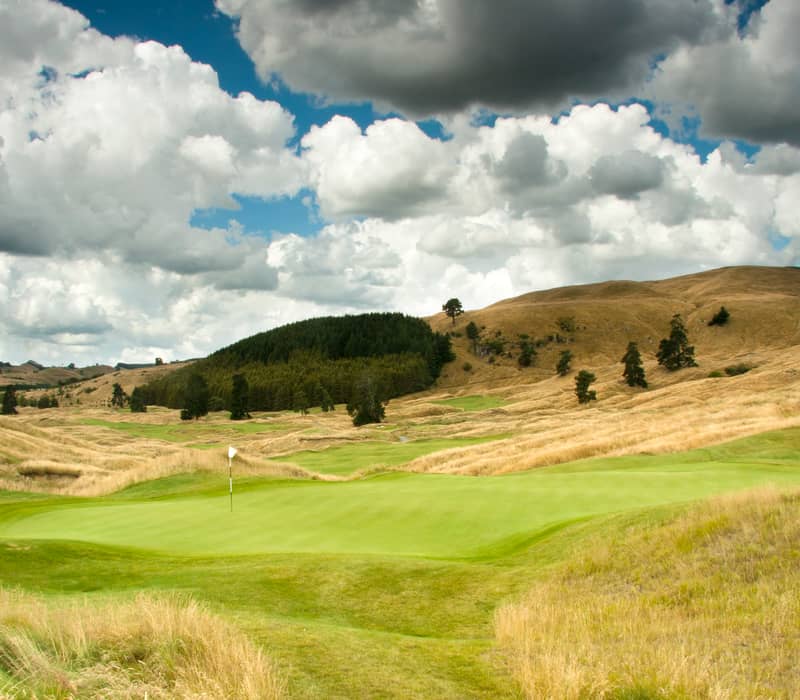 Golf course green with flag surrounded by hills and trees under a cloudy sky. Credit: Kinloch Golf