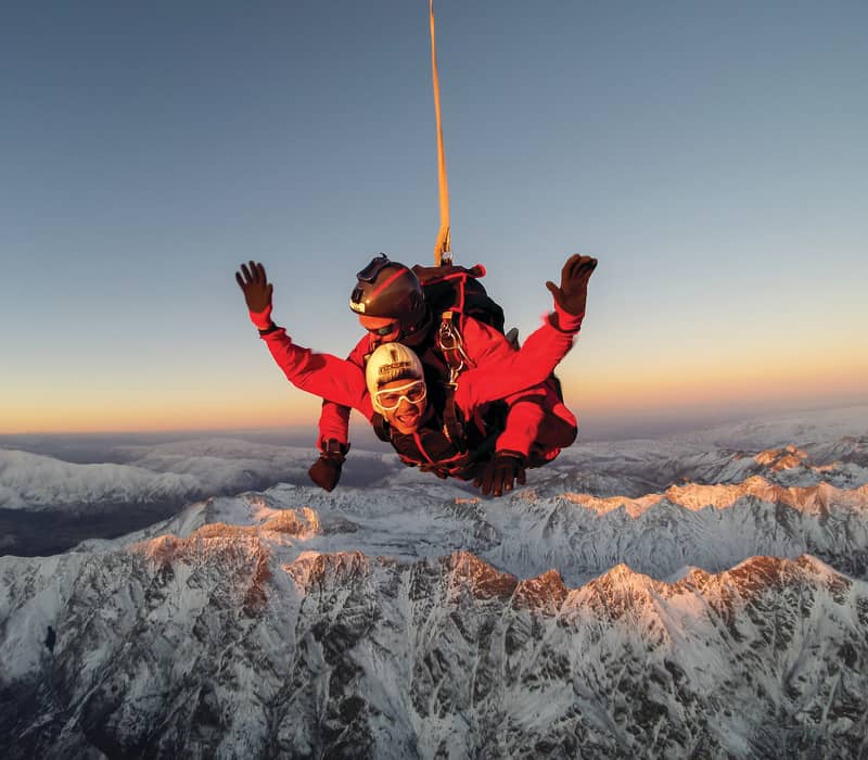 Two people tandem skydiving over snowy mountains at sunset